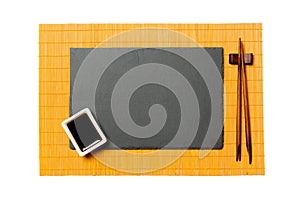 Empty rectangular black slate plate with chopsticks for sushi and soy sauce on yellow bamboo mat background. Top view with copy