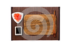 Empty rectangular brown wooden plate with chopsticks for sushi, ginger and soy sauce on dark bamboo mat background. Top view with