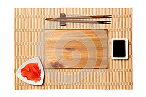Empty rectangular brown wooden plate with chopsticks for sushi, ginger and soy sauce on yellow bamboo mat background. Top view