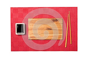 Empty rectangular brown wooden plate with chopsticks for sushi and soy sauce on red mat sushi background. Top view with copy space
