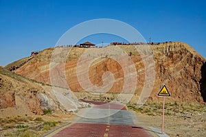 Empty road runs past a cliff with tourists standing on top of the colorful hill.