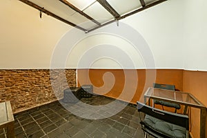 An almost empty room with black tile soils, brick wall and a metal table with two folding chairs and black square puffs