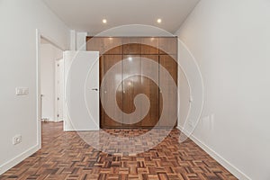 An empty room with a large built-in wardrobe with sapelly-colored wooden doors covering one wall and reddish parquet floors