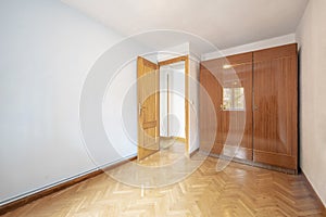 An empty room with a wardrobe with three-section glossy veneered doors with oak parquet floors placed in a herringbone pattern and