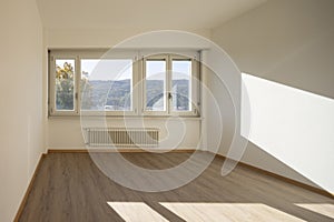 Empty room with white walls and a large, bright window that lets in direct sunlight. Outside the window are trees and hills