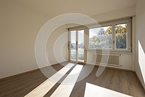 Empty room with white walls and a large, bright window that lets in direct sunlight. There is a radiator under the window. Outside