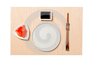 Empty round white plate with chopsticks for sushi, ginger and soy sauce on brown sushi mat background. Top view with copy space