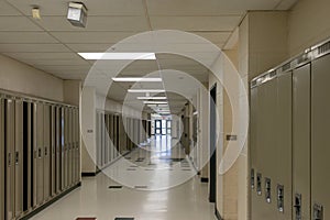 Empty school hallway - lined with closed lockers - beige walls - tiled floor - ceiling lights illuminating space - exit doors at