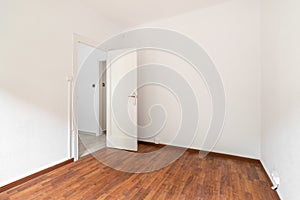 Empty spacious room well lit by daylight. The floor is dark brown wood parquet. White walls with a doorway opening onto