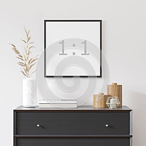 Empty square frame on white wall with black dresser, white vase and wood decor.