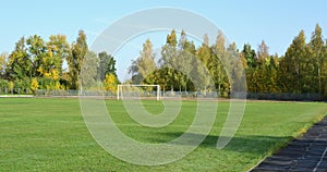 In an empty stadium, a green football field and white gates against the backdrop of an autumn forest and blue sky