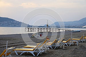 Empty sun loungers on a sandy beach against the backdrop of a pier, sea and mountains, evening, calm sea, deserted