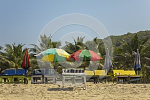 Empty sun loungers under the multi-colored beach umbrellas on the yellow sand against the green palm trees under a clear blue sky