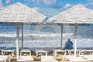 Empty sun loungers under umbrellas on seashore in storm. A lonely man sits with his back and looks at the waves