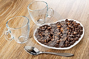 Empty transparent cups, roasted coffee beans in saucer in heart shape, spoon on wooden table