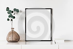 Empty vertical frame mockup in modern minimalist interior with plant in trendy vase on white wall background.