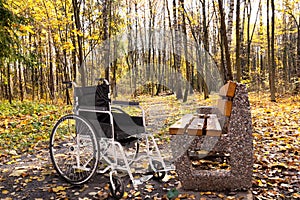 An empty wheelchair parked in an autumn park next to a bench. The concept of accessibility of people with disabilities