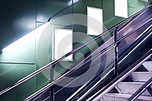 Empty white advertising urban billboard indoor in subway hall, vertical portrait, green walls, above metallic stairs.