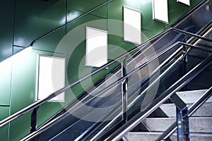 Empty white advertising urban billboard indoor in subway hall, vertical portrait, green walls, above metallic stairs.