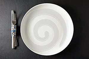 Empty white ceramic plate with cutlery wrapped in hundred dollar bill on black stone table. Top view with copy space