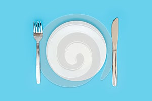 Empty white ceramic plate with a fork and a table knife on a blue background, top view