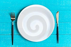 Empty white ceramic round plate with knife and fork on blue wooden background