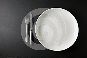 Empty white dinner plate and cutlery on a black stone table. Top view with copy space