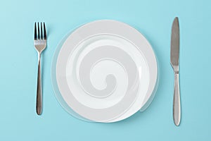 Empty white dinner plate, table knife and fork on a blue background. Set table. Copy space on dish for food design