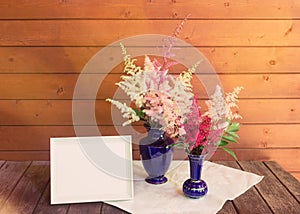 Empty white frame and red, white and pink beautiful astilbe flowers in blue vases on wooden table. Selective focus