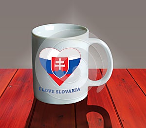 Prázdný bílý hrnek na dřevěném stole s vlajkou a státním znakem Slovenska v podobě srdce a nápisem I love
