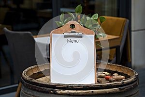 Empty white paper menu on restaurant entrance. Modern and cozy cafe or bar facade with tables and chairs on outdoor patio.