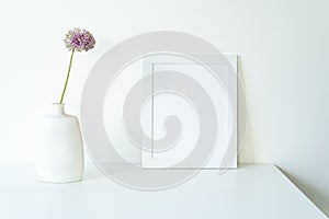 Empty white picture frame mockup. Modern and elegant vase with allium ampeloprasum flower over white table with white background.