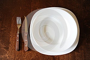 Empty white plate with a fork and knife on a dark wooden brown table
