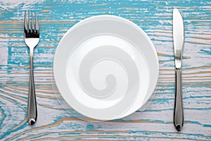 Empty white plate with silver fork and knife on blue wooden table. Dinner place setting. Top view