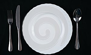 Empty white plate with silver fork, knife and spoon isolated on black background. Dinner place setting. Top view.