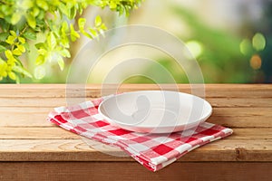 Empty white plate on wooden table with red checked tablecloth over  blurred nature background.  Kitchen mock up for design and