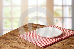 Empty white plate on wooden table with red checked tablecloth over  blurred window background.  Kitchen mock up for design and
