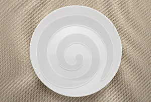 Empty white plate on woven napkin. Clean plate on light brown tablecloth. Unused dishes.