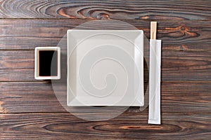 Empty white square plate with chopsticks for sushi and soy sauce on wooden background. Top view with copy space