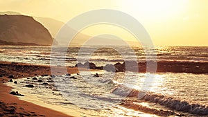 Empty wild beach lit by golden sunset. Small waves over rock, hills in hazy background