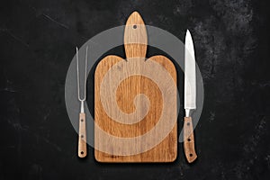 Empty wooden cutting board, meat fork and knife on black stone background. Top view, flat lay, copy space. Place for your design