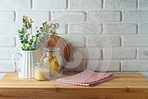 Empty wooden l table with tablecloth, plant, food jars and cutting board over white brick wall background. Kitchen mock up for