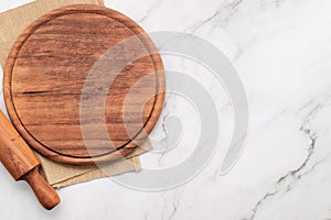 Empty wooden pizza platter with napkin and rolling pin set up on marble stone kitchen table. Pizza board and tablecloth on white