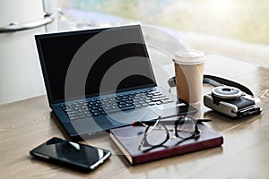 Empty workplace. Near window on desk is laptop, smartphone, notebook, glasses, cup of coffee, camera for instant photos.