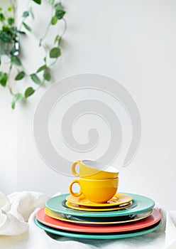 Empty yellow cups and brightly colored plates on a white table covered with a linen tablecloth, a flower pot on the wall in the