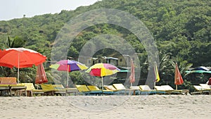 Empty yellow sun loungers under multi-colored beach umbrellas on the wind on the yellow sand against the background of a green mou