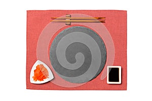 Emptyround black slate plate with chopsticks for sushi, ginger and soy sauce on red napkin background. Top view with copy space