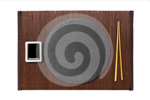 Emptyround black slate plate with chopsticks for sushi and soy sauce on dark bamboo mat background. Top view with copy space for
