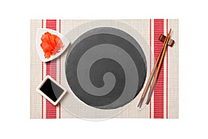 Emptyround black slate plate with chopsticks for sushi and soy sauce, ginger on sushi mat background. Top view with copy space for