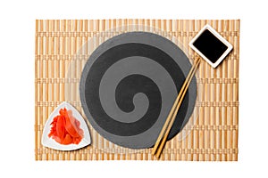 Emptyround black slate plate with chopsticks for sushi and soy sauce, ginger on yellow bamboo mat background. Top view with copy